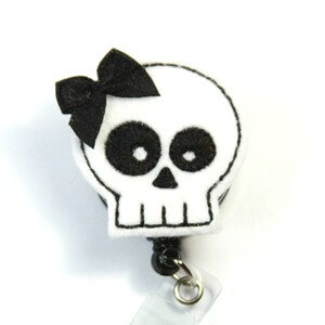 May include: White felt skull with black embroidered eyes and a black felt bow. The skull is attached to a black retractable badge reel with a clear plastic badge holder.