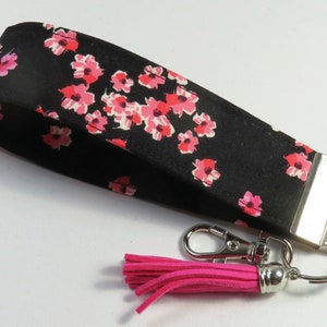 May include: A black fabric key chain with a pink floral pattern and a pink tassel. The key chain has a silver metal ring and a silver metal loop.