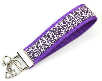 May include: Purple key chain with a white and black floral pattern. The key chain has a silver metal key ring.