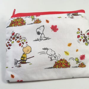 May include: White zippered pouch featuring a fall-themed print with Snoopy and Charlie Brown playing in leaves. The pouch has a red zipper and a white strap with a metal ring closure. The design includes red, yellow, and orange leaves.