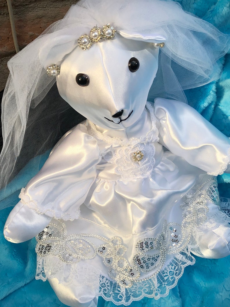 K&ouml;nnte beinhalten: Wei&szlig;er Satin-Teddyb&auml;r, der ein wei&szlig;es Brautkleid mit Schleier, Spitzenbesatz und Perlenverzierungen tr&auml;gt.