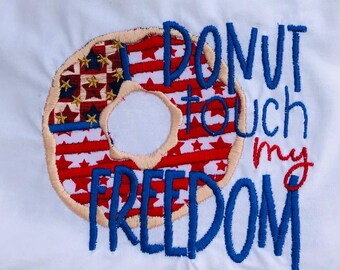 Könnte beinhalten: Ein weißes T-Shirt mit einem blauen und roten gestickten Donut mit Sternen und Streifen. Der Text "Donut touch my freedom" ist in Blau unter dem Donut gestickt.