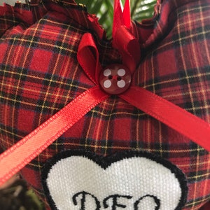 May include: Red and black plaid heart-shaped pillow with a white heart patch that says "DEO" in black script. The pillow has a red ribbon tied around it with a red button in the center.