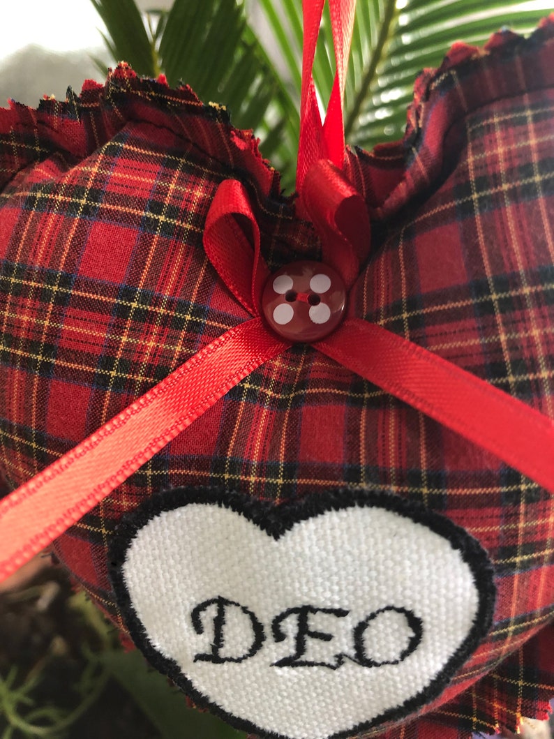 May include: Red and black plaid heart-shaped pillow with a white heart patch that says "DEO" in black script. The pillow has a red ribbon tied around it with a red button in the center.