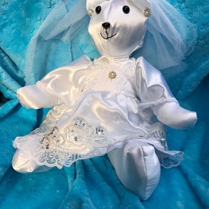 K&ouml;nnte beinhalten: Wei&szlig;er Satin-Teddyb&auml;r, der ein wei&szlig;es Satin-Kleid mit Spitzenbesatz und Schleier tr&auml;gt. Der B&auml;r hat eine Tiara und Perlenohrringe. Der B&auml;r liegt auf einer blauen flauschigen Decke.