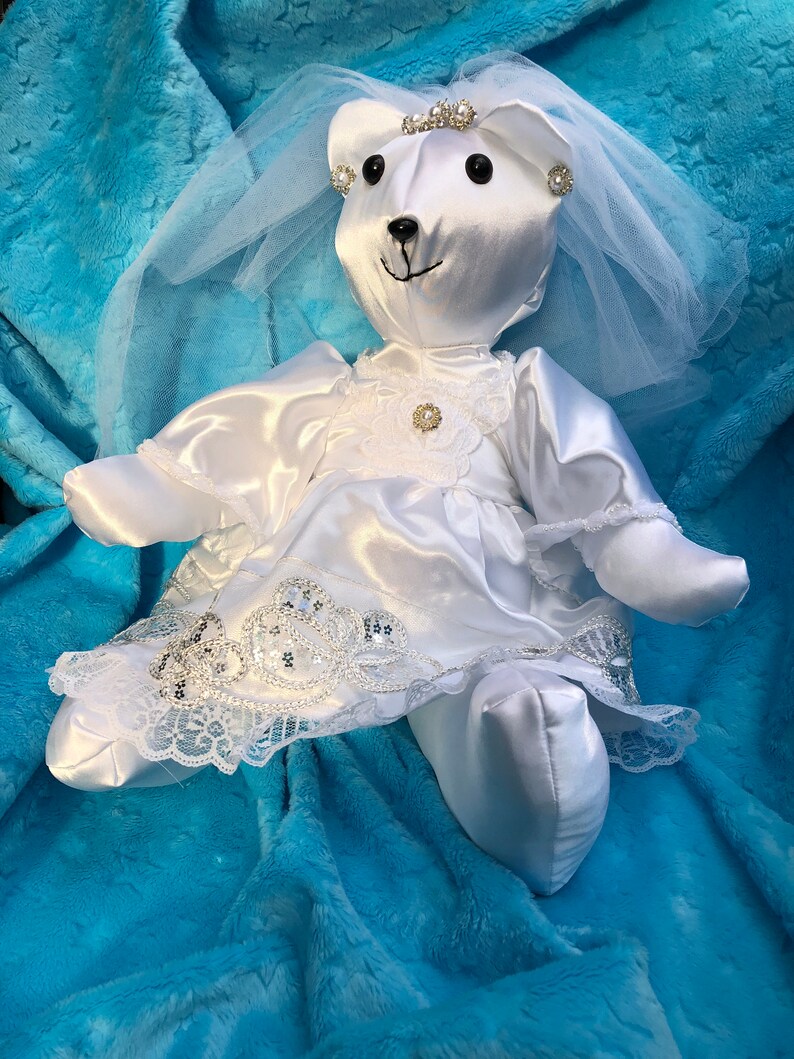 K&ouml;nnte beinhalten: Wei&szlig;er Satin-Teddyb&auml;r, der ein wei&szlig;es Satin-Kleid mit Spitzenbesatz und Schleier tr&auml;gt. Der B&auml;r hat eine Tiara und Perlenohrringe. Der B&auml;r liegt auf einer blauen flauschigen Decke.