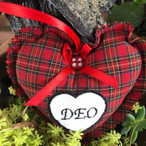 May include: Red and black plaid heart-shaped pillow with a white heart embroidered with the word "DEO" and a red button with white dots in the center.