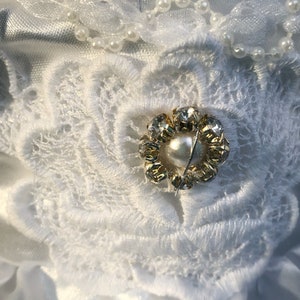 K&ouml;nnte beinhalten: Ein wei&szlig;er Satinstoff mit einer wei&szlig;en Spitzenblumenverzierung. Die Blume hat eine goldene und klare Strassmitte mit einer Perle in der Mitte.