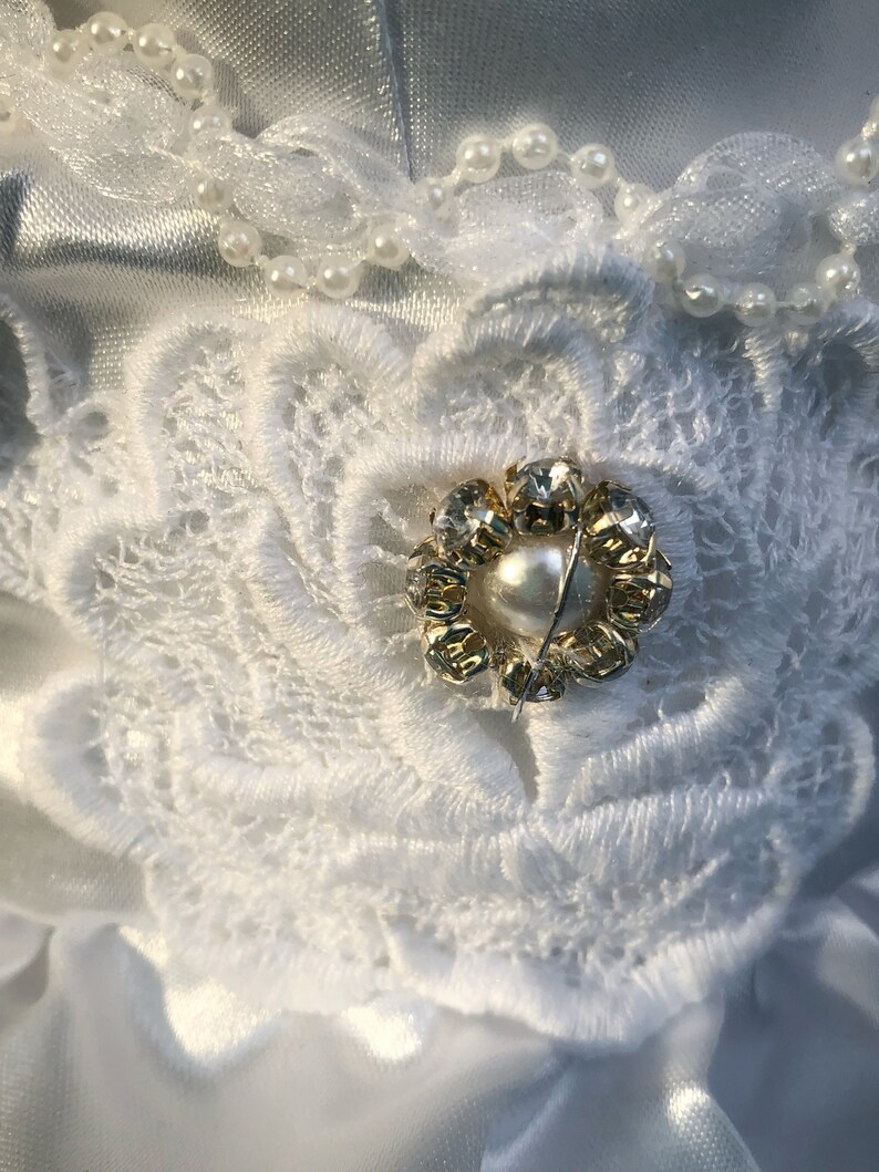 K&ouml;nnte beinhalten: Ein wei&szlig;er Satinstoff mit einer wei&szlig;en Spitzenblumenverzierung. Die Blume hat eine goldene und klare Strassmitte mit einer Perle in der Mitte.