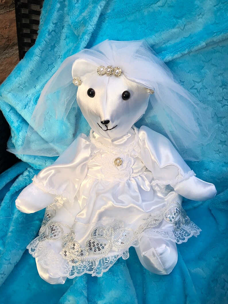 K&ouml;nnte beinhalten: Ein wei&szlig;er Satin-Teddyb&auml;r, der ein wei&szlig;es Satin-Hochzeitskleid mit einer Spitzenborte und einem T&uuml;llschleier tr&auml;gt. Der B&auml;r tr&auml;gt eine Perlenkette und Ohrringe.