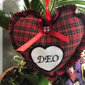 May include: Red and black plaid heart-shaped pillow with a white heart embroidered with the word "DEO" and a red ribbon with a white button.