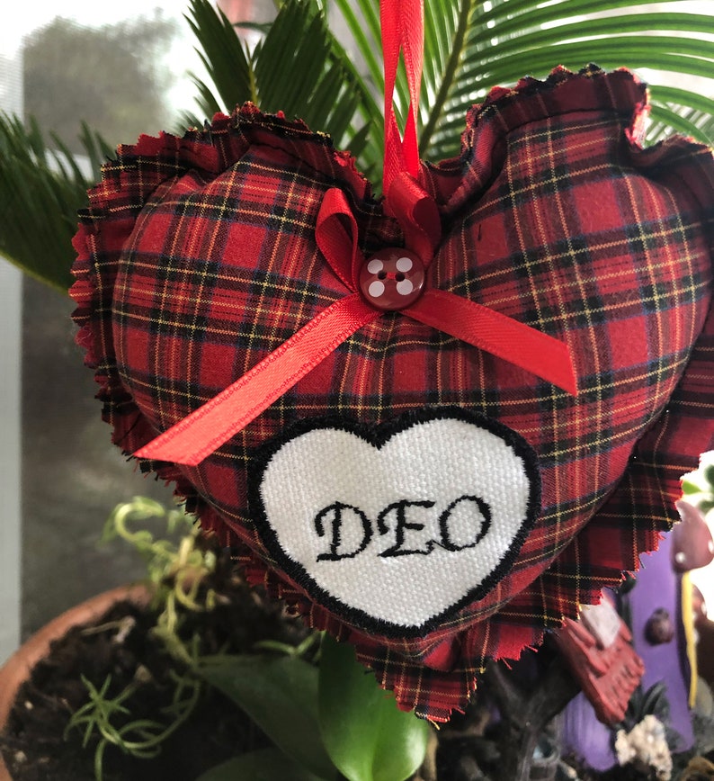 May include: Red and black plaid heart-shaped pillow with a white heart embroidered with the word "DEO" and a red ribbon with a white button.
