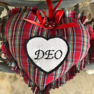 May include: Red and green plaid heart-shaped pillow with a white heart-shaped patch that says "DEO" in black embroidery. The pillow has a red ribbon with white polka dots and is attached to a red ribbon.