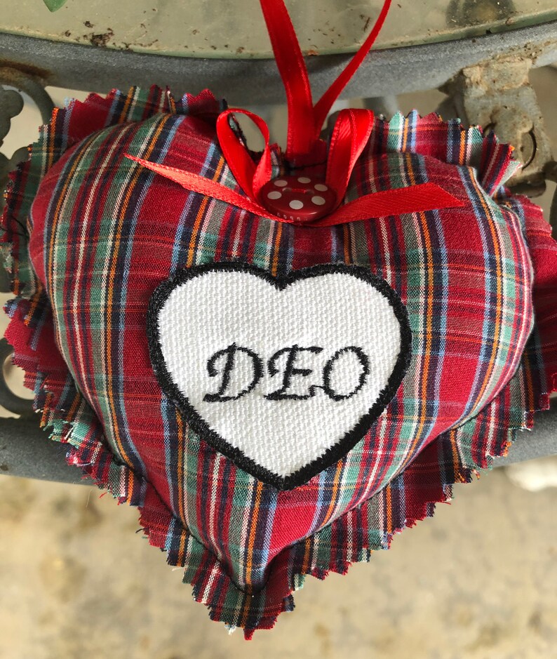 May include: Red and green plaid heart-shaped pillow with a white heart-shaped patch that says "DEO" in black embroidery. The pillow has a red ribbon with white polka dots and is attached to a red ribbon.