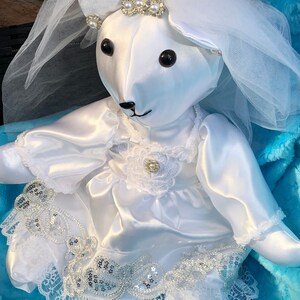 K&ouml;nnte beinhalten: Ein wei&szlig;er Satin-Teddyb&auml;r, der ein wei&szlig;es Satin-Kleid mit Spitzenbesatz und einen wei&szlig;en T&uuml;llschleier tr&auml;gt. Der B&auml;r hat schwarze Augen und eine schwarze Nase. Der B&auml;r tr&auml;gt eine Perlen- und Strass-Tiara.
