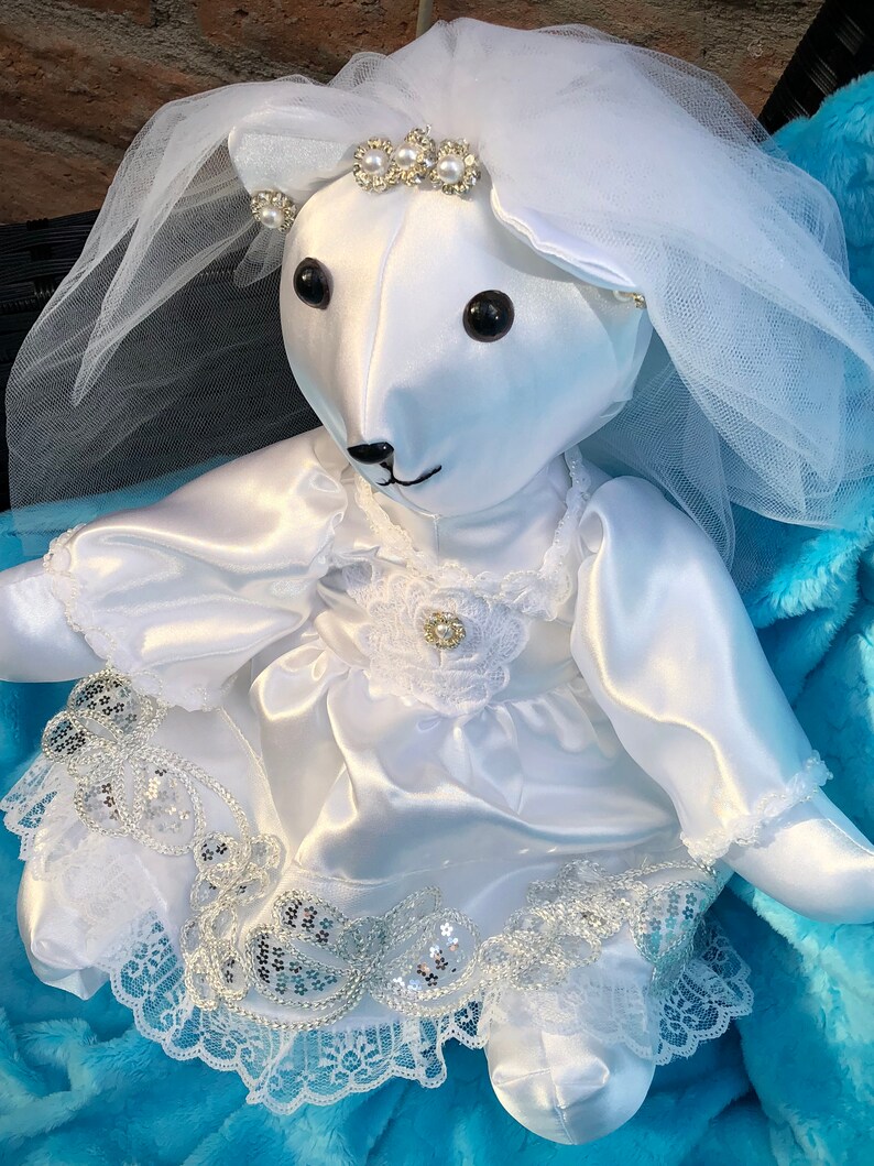 K&ouml;nnte beinhalten: Ein wei&szlig;er Satin-Teddyb&auml;r, der ein wei&szlig;es Satin-Kleid mit Spitzenbesatz und einen wei&szlig;en T&uuml;llschleier tr&auml;gt. Der B&auml;r hat schwarze Augen und eine schwarze Nase. Der B&auml;r tr&auml;gt eine Perlen- und Strass-Tiara.