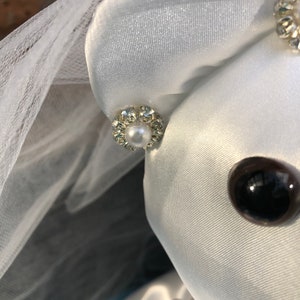 K&ouml;nnte beinhalten: Ein wei&szlig;er Satin-Teddyb&auml;r, der einen wei&szlig;en Schleier und einen Perlen- und Strassohrring tr&auml;gt. Der B&auml;r hat ein schwarzes Knopfauge.