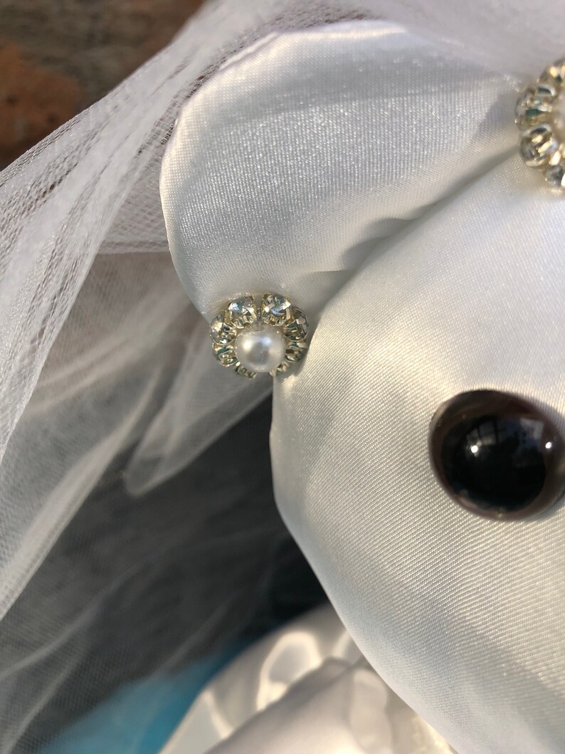 K&ouml;nnte beinhalten: Ein wei&szlig;er Satin-Teddyb&auml;r, der einen wei&szlig;en Schleier und einen Perlen- und Strassohrring tr&auml;gt. Der B&auml;r hat ein schwarzes Knopfauge.