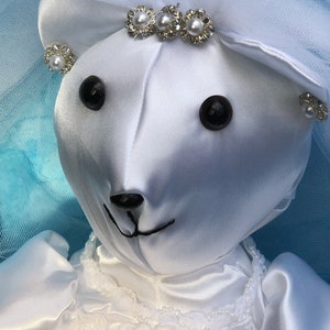 K&ouml;nnte beinhalten: Wei&szlig;er Satin-Teddyb&auml;r, der ein wei&szlig;es Brautkleid mit Schleier und Perlenakzenten tr&auml;gt. Der B&auml;r hat schwarze Augen und eine schwarze Nase.