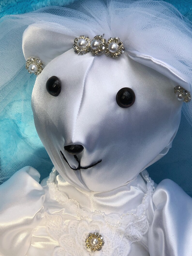K&ouml;nnte beinhalten: Wei&szlig;er Satin-Teddyb&auml;r, der ein wei&szlig;es Brautkleid mit Schleier und Perlenakzenten tr&auml;gt. Der B&auml;r hat schwarze Augen und eine schwarze Nase.