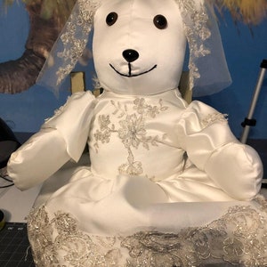 K&ouml;nnte beinhalten: Ein wei&szlig;er Teddyb&auml;r, der ein wei&szlig;es Satin-Hochzeitskleid mit Spitzenbesatz und Schleier tr&auml;gt. Der B&auml;r sitzt auf einem Stuhl.