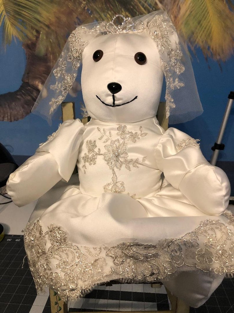 K&ouml;nnte beinhalten: Ein wei&szlig;er Teddyb&auml;r, der ein wei&szlig;es Satin-Hochzeitskleid mit Spitzenbesatz und Schleier tr&auml;gt. Der B&auml;r sitzt auf einem Stuhl.