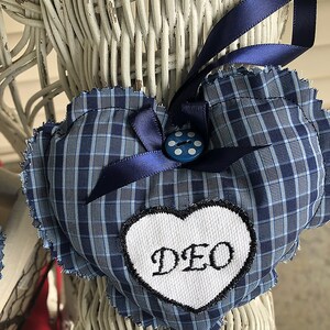 May include: Blue and white plaid heart-shaped pillow with a white heart-shaped patch that says "DEO" in black embroidery. The pillow has a blue ribbon and a blue button with white dots.
