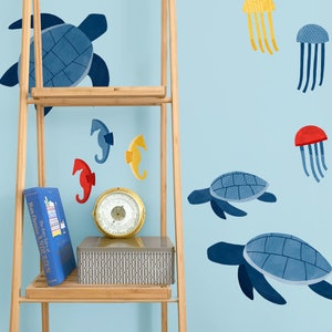 Puede incluir: Un conjunto de pegatinas de pared de tortugas marinas azules y blancas, caballitos de mar rojos y amarillos y medusas rojas y azules. Las pegatinas están en una pared azul claro con un estante de bambú y un libro titulado "El cuento de hadas de Hans Christian Andersen".
