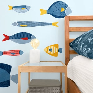 Può includere: Un muro blu chiaro con adesivi di pesci colorati. Gli adesivi sono di diverse dimensioni e forme, tra cui un pesce lungo e sottile, un pesce rotondo e un pesce con una coda lunga. Il muro è anche decorato con una struttura del letto in legno e un piccolo tavolino da comodino in legno.