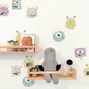 May include: A white wall with colorful monster stickers. A gray knitted stuffed toy sits on a wooden shelf with a baby monitor and a book titled "Brown Bear, Brown Bear, What Do You See?" by Bill Martin Jr. and Eric Carle.