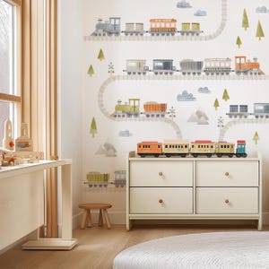 May include: A child's room with a train theme. The wall features a painted mural of toy trains on tracks, clouds, and trees. A white dresser with wooden train toys on top sits below the mural. A small wooden stool and a round rug complete the scene.