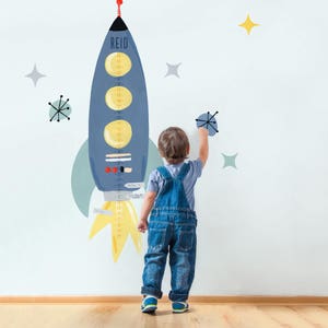 May include: A blue and yellow rocket ship height chart with a child standing next to it. The chart has the name "Reid" at the top and a scale in inches. The child is wearing a blue shirt and overalls.