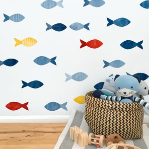 Pode incluir: Uma cesta de vime cheia de animais de pelúcia fica em frente a uma parede com adesivos de peixes azuis, vermelhos e amarelos. A parede é pintada de azul claro.