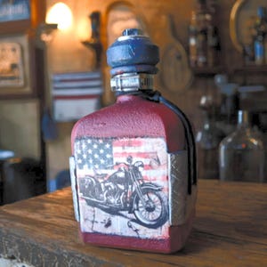 May include: A red canteen with a textured finish featuring a graphic of a motorcycle against an American flag background. The canteen has a blue cap, a metal hose clamp around the neck, and silver diamond-plate accents on the sides.