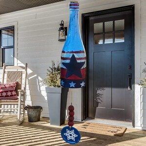 May include: A patriotic wind chime shaped like a bottle, painted blue, red, and white with a large black star and white stars. The wind chime hangs from a string with red beads and a blue, star-shaped pendant. The background includes a white house and a black door.