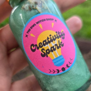 May include: A green glass bottle with a gold lid and a pink and blue label that says "Creativity Spark" with a lightbulb graphic. The label also has the text "The Owl Witch Shop" and a crescent moon graphic.