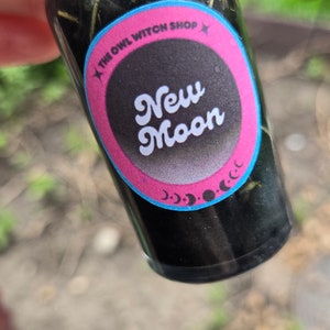 May include: A small glass bottle filled with a dark liquid and herbs. The bottle has a pink and blue label that says "The Owl Witch Shop" and "New Moon".
