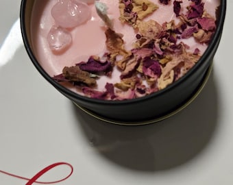 May include: A pink candle with rose petals and clear quartz crystals in a black tin container. The candle is on a white plate with the word "love" written in red.