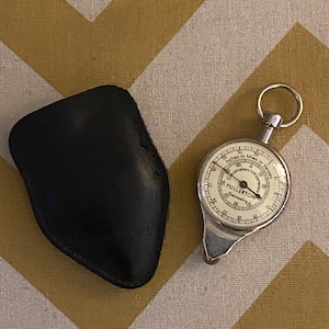 May include: A vintage Fullerton map measurer with a silver-colored body and a white dial, next to a black leather case. The dial reads "Inches to Miles or Yards" and "Fullerton Germany."