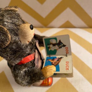 May include: A vintage wind-up toy bear wearing a red strap and holding a small book with pictures of a dog and a duck. The book says "DOG" and "DUCK" on the pages.
