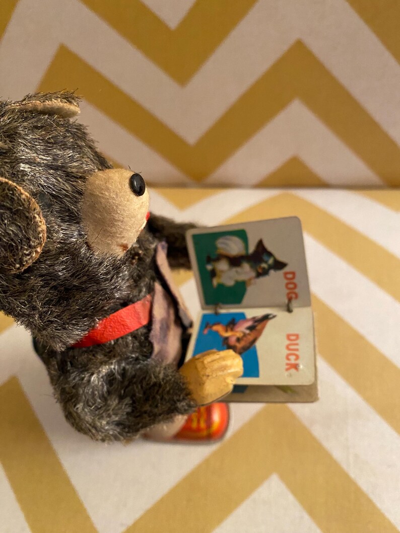 May include: A vintage wind-up toy bear wearing a red strap and holding a small book with pictures of a dog and a duck. The book says "DOG" and "DUCK" on the pages.