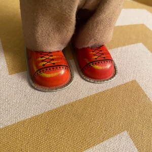 May include: Close-up of a pair of red shoes with black laces and yellow accents. The shoes are made of metal and have a silver trim. The shoes are worn by a vintage tin toy.