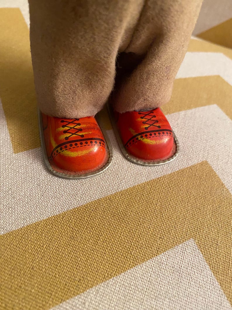 May include: Close-up of a pair of red shoes with black laces and yellow accents. The shoes are made of metal and have a silver trim. The shoes are worn by a vintage tin toy.