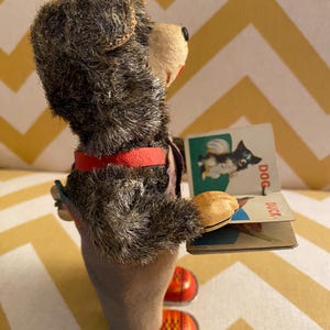 May include: A vintage wind-up toy bear wearing overalls and red shoes holds a book with pictures of a dog and a duck. The book has the words "DOG" and "DUCK" printed on it.