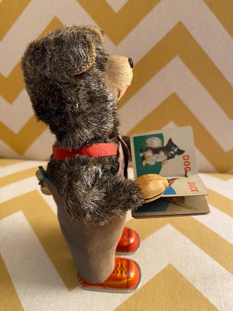 May include: A vintage wind-up toy bear wearing overalls and red shoes holds a book with pictures of a dog and a duck. The book has the words "DOG" and "DUCK" printed on it.