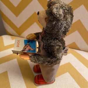 May include: A vintage wind-up toy bear wearing overalls and red shoes. The bear is holding a card that says "Duck" and has a picture of a duck on it.