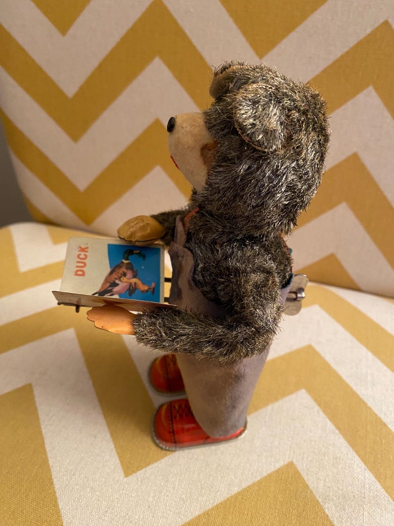 May include: A vintage wind-up toy bear wearing overalls and red shoes. The bear is holding a card that says "Duck" and has a picture of a duck on it.