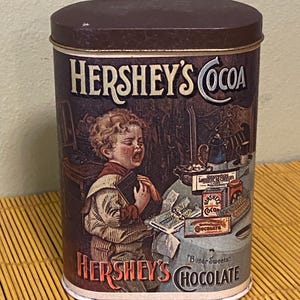 May include: Vintage Hershey's Cocoa tin with a dark brown lid and a rectangular shape. The tin features an illustration of a child and the words "Hershey's Cocoa" and "Bitter Sweets Chocolate". The tin is a collectible item.