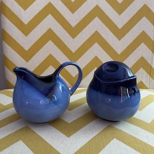 May include: A blue and white ceramic creamer and sugar bowl set. The creamer has a rounded body and a curved handle. The sugar bowl has a lid with a knob on top.
