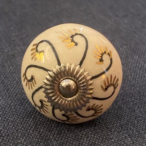 May include: A white ceramic knob with a gold and black floral design. The knob has a silver metal center.